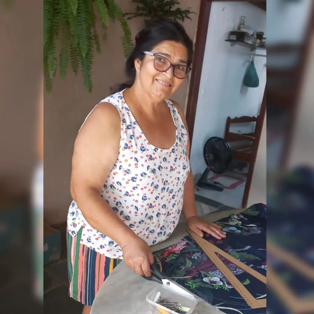 Maria Elena cortando um tecido estampado sobre a mesa do ateliê, com régua e alfinetes ao lado.